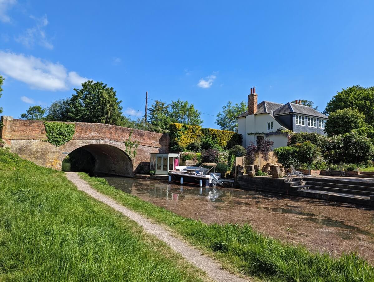 Sunny day next to the Basingstoek Canal, with a dirt path passing under a brick bridge with a two storey building on the other side of the water