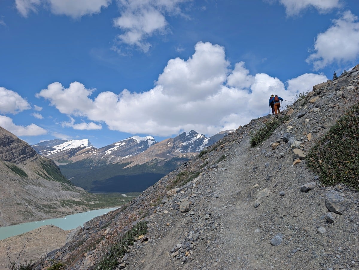 Looking up the narrow trail to two hikers above, with views of glacial lake and mountains below