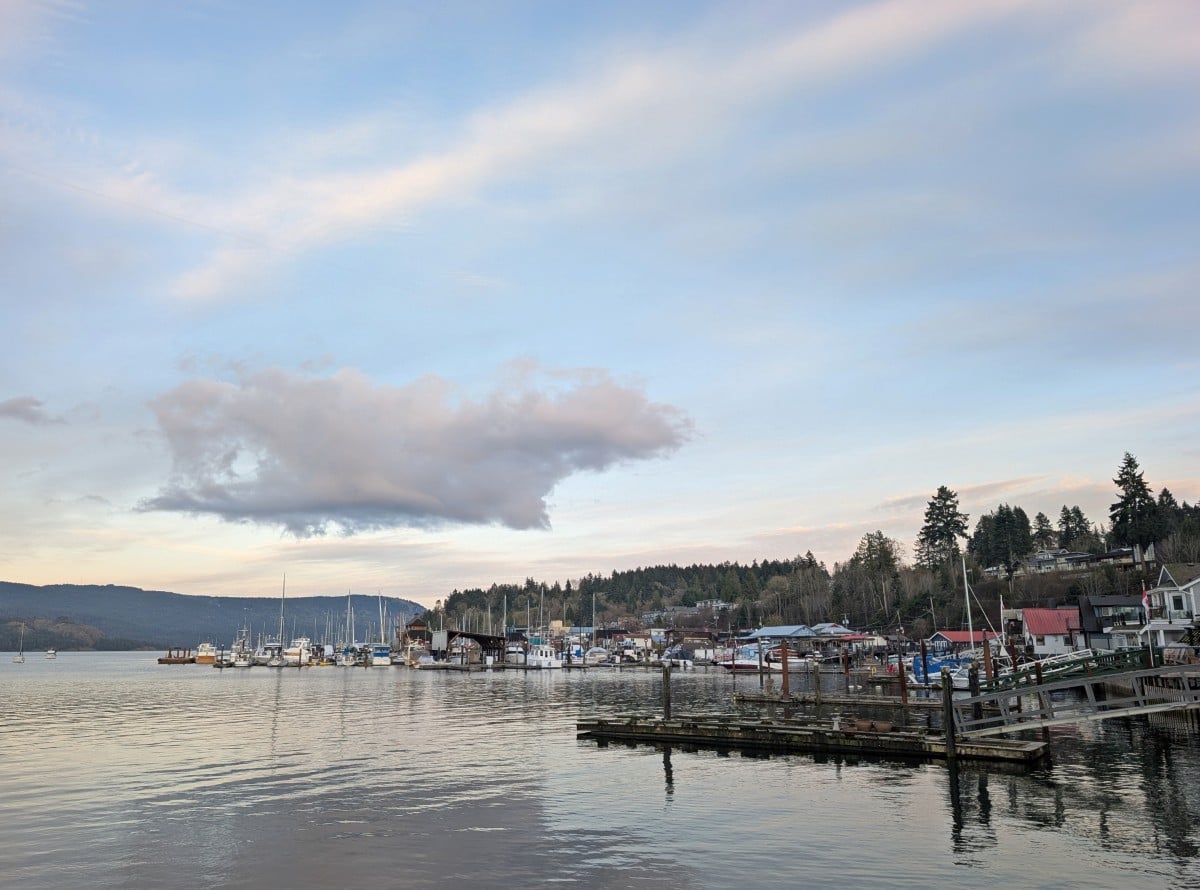 Cowichan Bay waterfront with calm ocean, lots of docks and wharves, buildings on stilts to the right and moored boats, around sunset