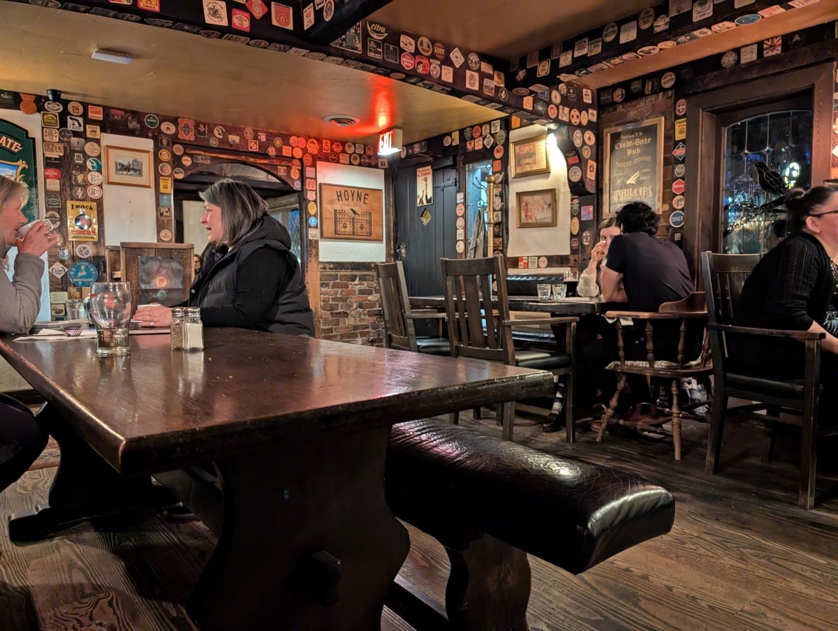 View of English style pub, the Crow & Gate, featuring large wooden tables, mismatched chairs, low wooden ceiling beams and pub paraphernalia all over the walls