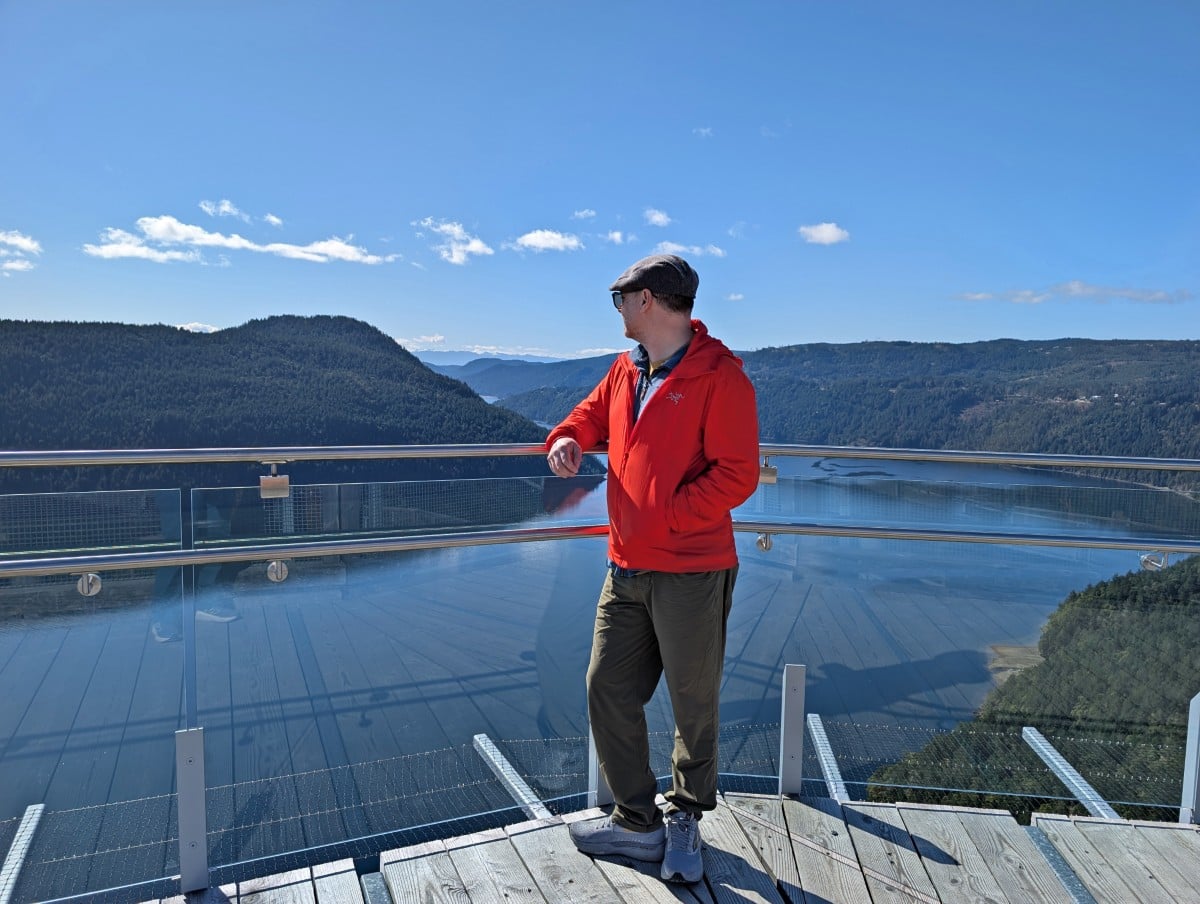 JR stands in a red jacket in front of a glass fence at the top of the Malahat Skywalk Spiral Tower, looking out to beautiful ocean inlet and peninsula views behind