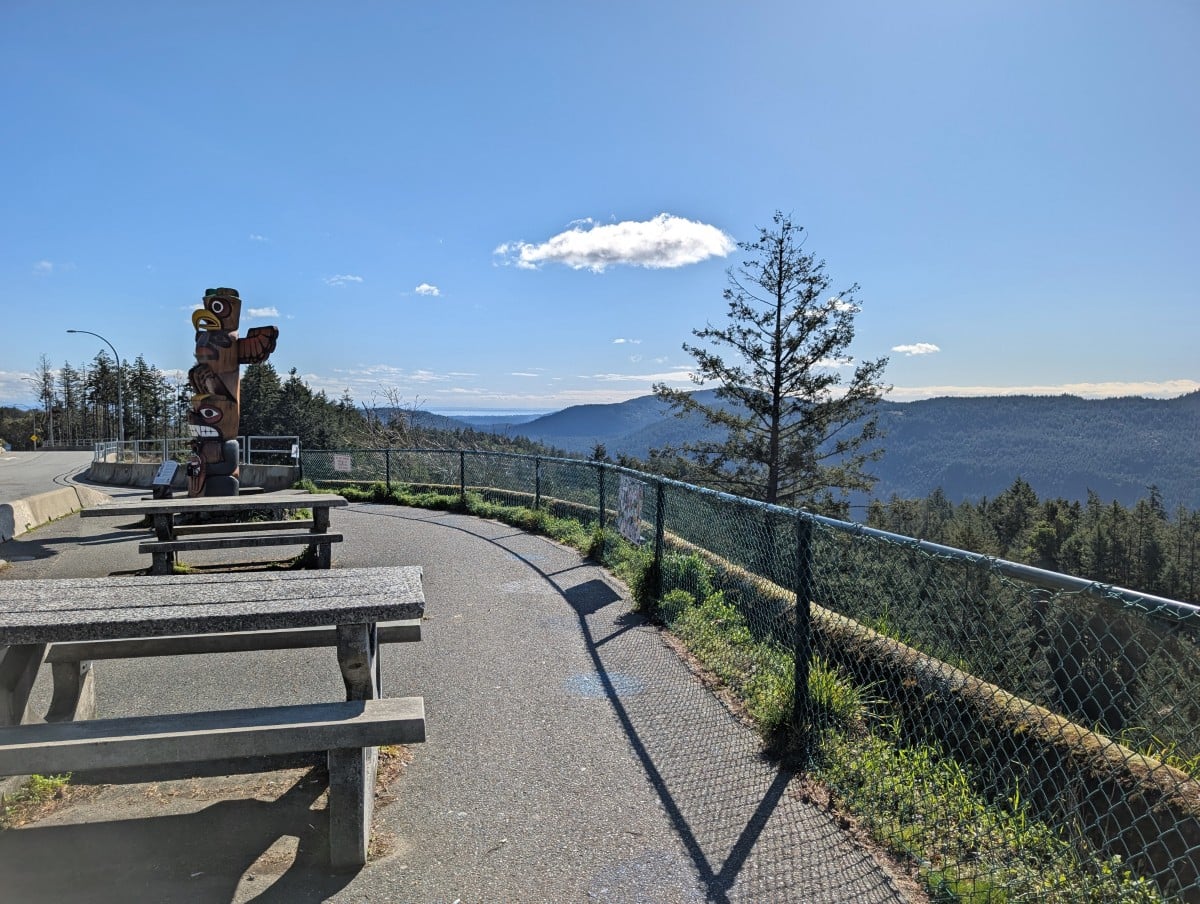 Side view of paved Malahat Summit viewpoint area with fencing, picnic tables and totem pole, overlooking elevated views of forest