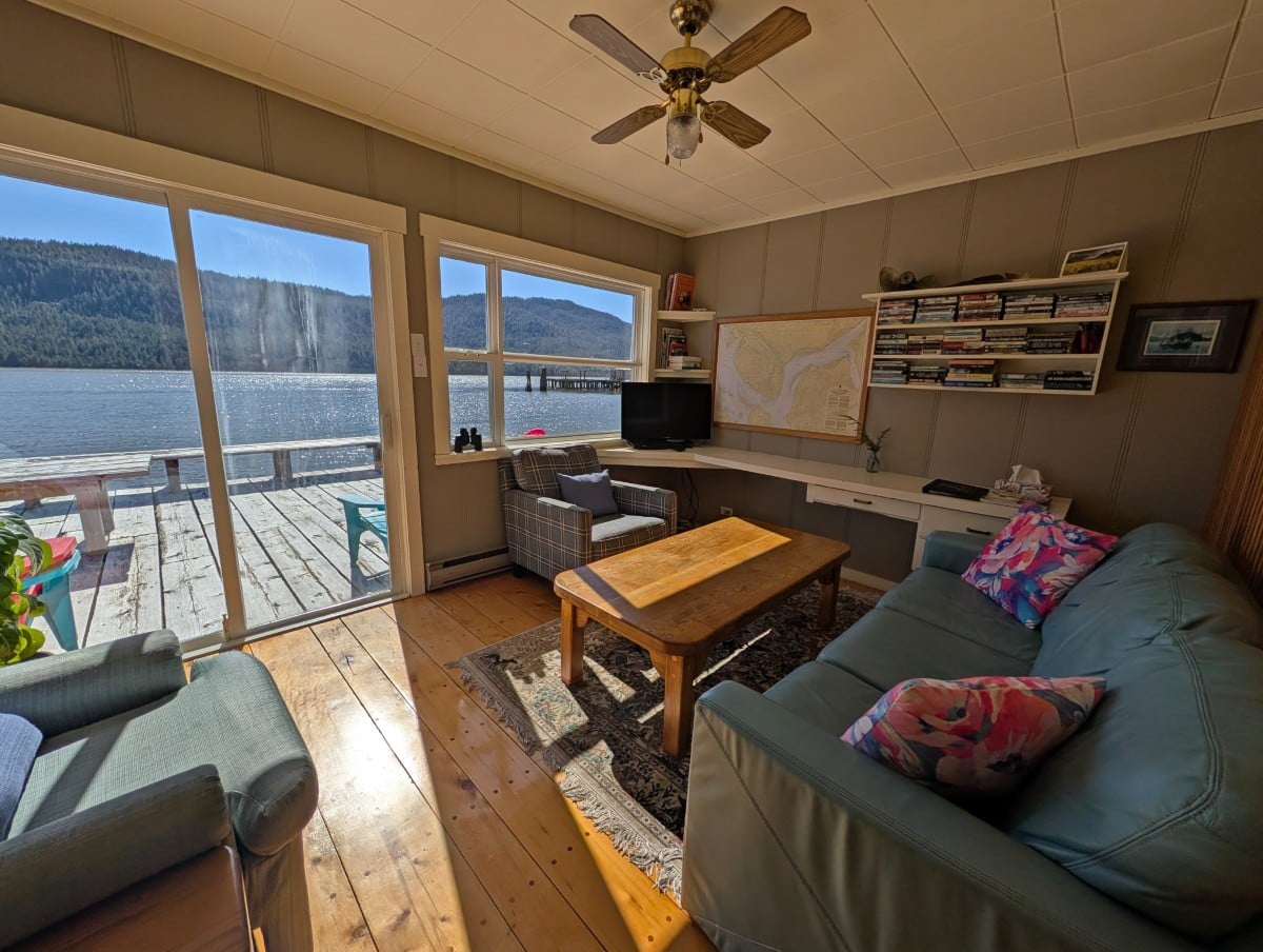 Wide angle view of Coho guesthouse living area, with couch, two chairs, bookcase and marine chart. A deck is visible outside the patio doors with ocean views