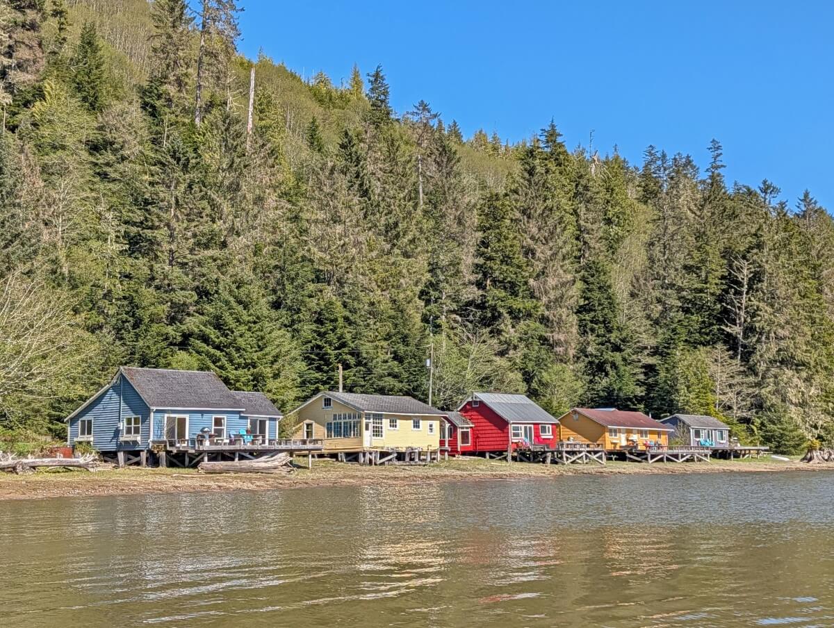Boat view of colourful cabins at the Cassiar Cannery that sit on the edge of the Skeena River estuary, a forested slope behind them 