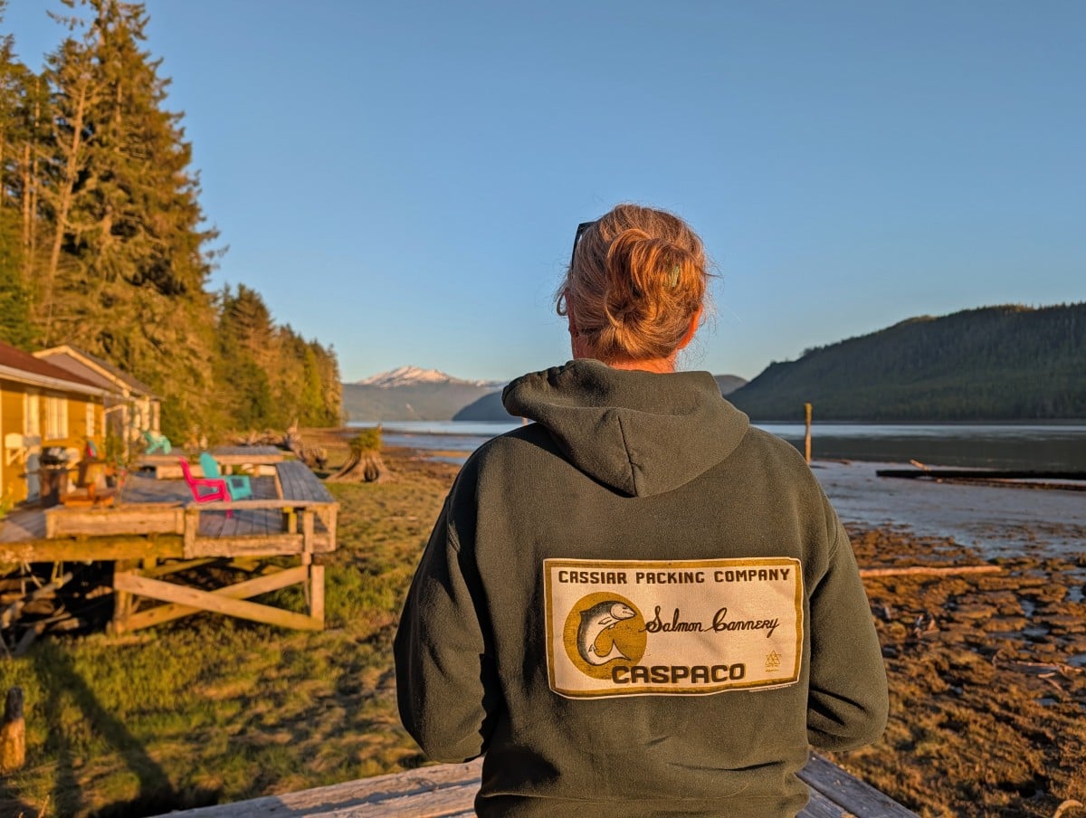 Back view of woman wearing Cassiar Packing Company logo hoody, with view of cottage decks and ocean behind