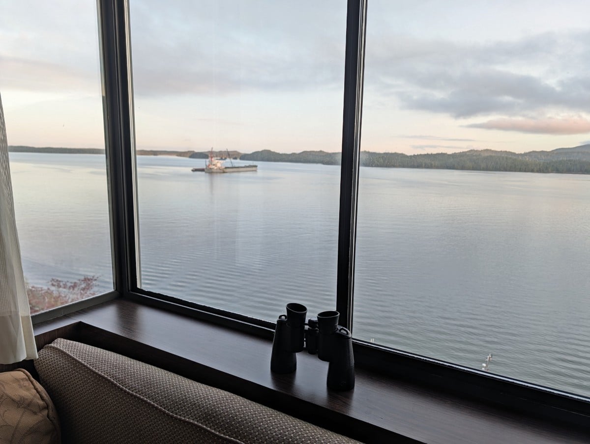 Crest Hotel room view, with window seat visible, below window shelf that has a pair of binoculars . Outside, the ocean is calm, with sunrise colours and a large boat