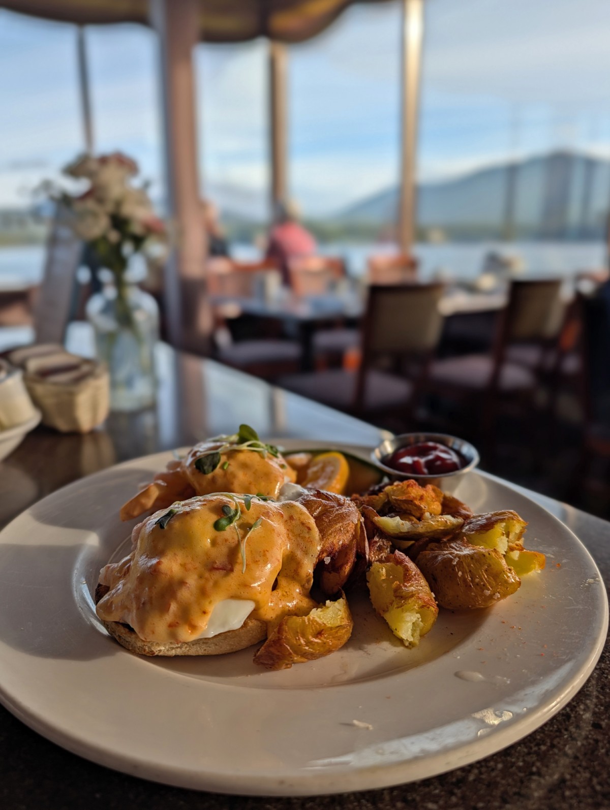 Above front view of Eggs Benedict dish on white plate at Waterfront Restaurant at the Crest Hotel in Prince Rupert, with seating and ocean views in the background
