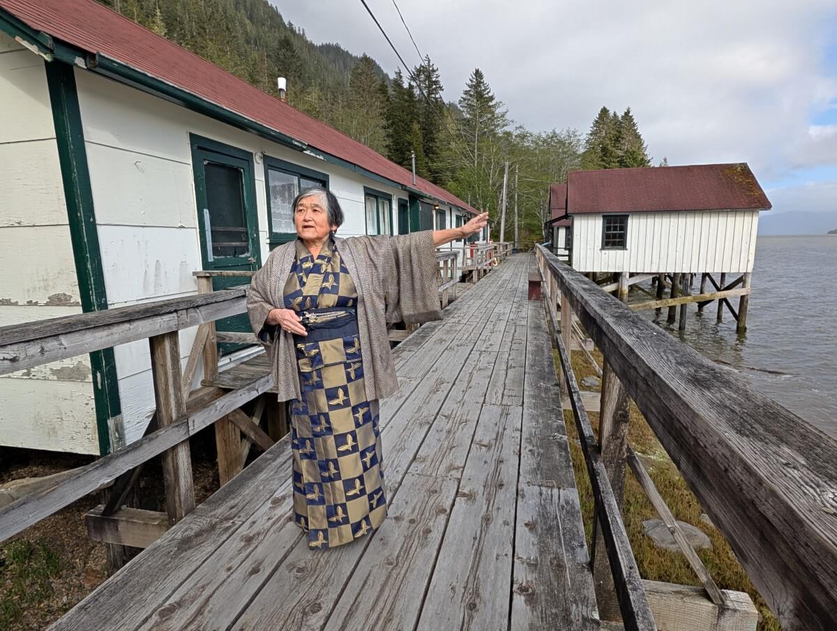 Mona Izumi stands wearing a kimono, gesticulating towards the ocean while standing on the boardwalk at the North Pacific Cannery, with Japanese section buildings behind