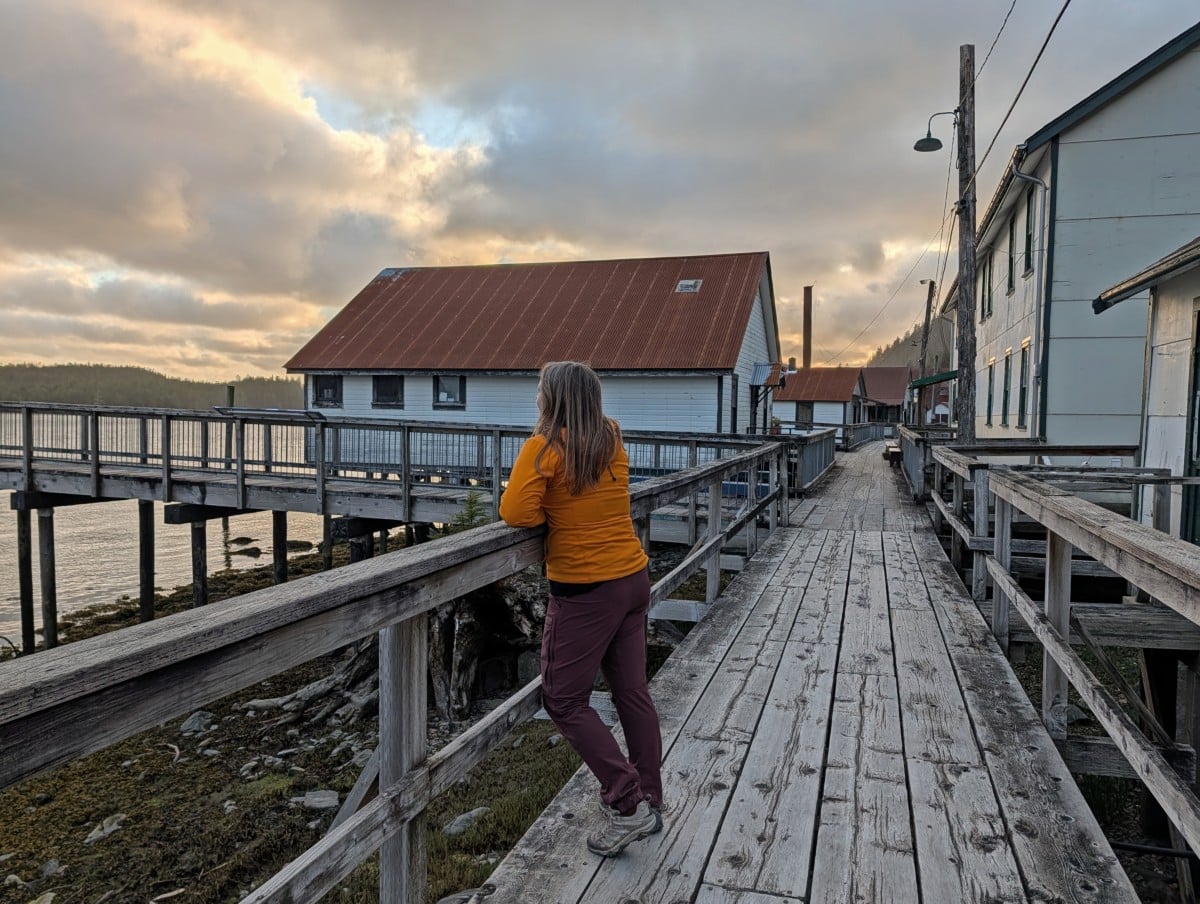 Back view of Gwmma standing on the boardwalk at North Pacific Cannery, looking out to the ocean. The boardwalk stretches into the distance, with small buildings on either side