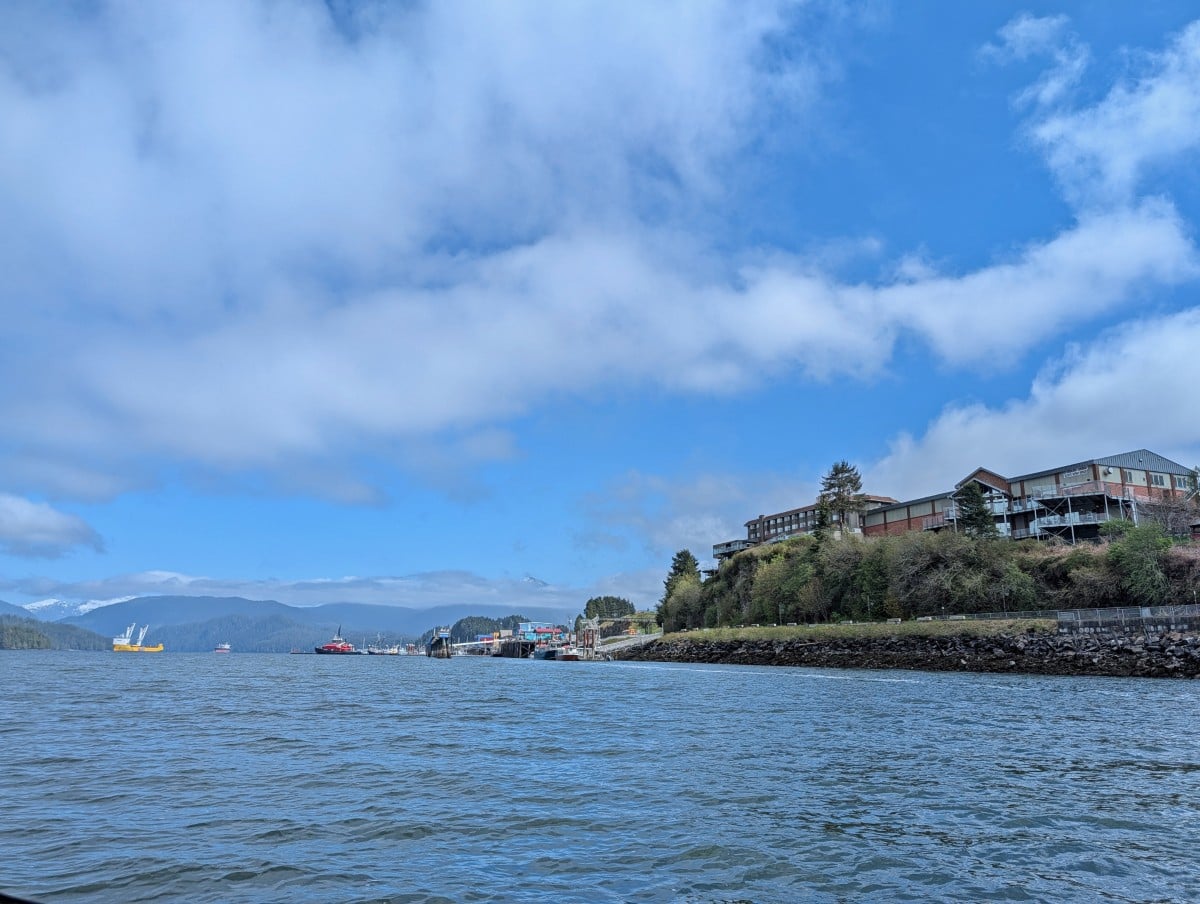 Boat view of Prince Rupert waterfront, with sea level walkway and hotels perched on top of a hill. Many boats are visible in the background as well as mountains