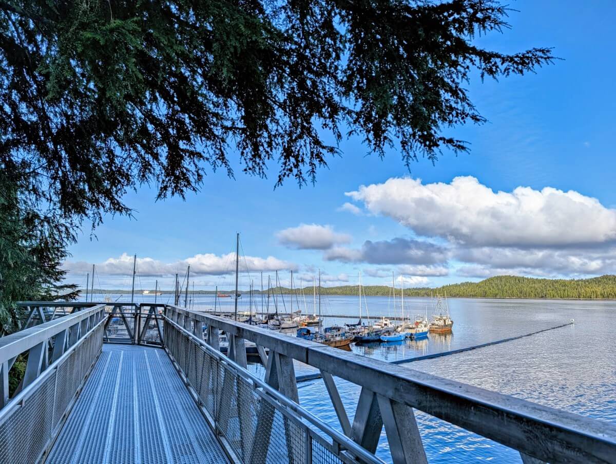 A metal bridge along the coastline in Prince Rupert, part of the Rushbrook Trail. The ocean is calm below the bridge, with boats in the marina and forested hills in the background