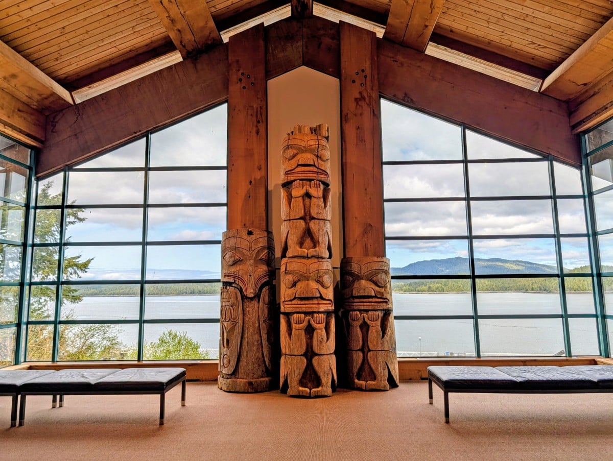 Monumental gallery at the museum with floor to ceiling windows with three totem poles in the middle, bench seating in front. Outside of the window, the ocean and forested hills are visible