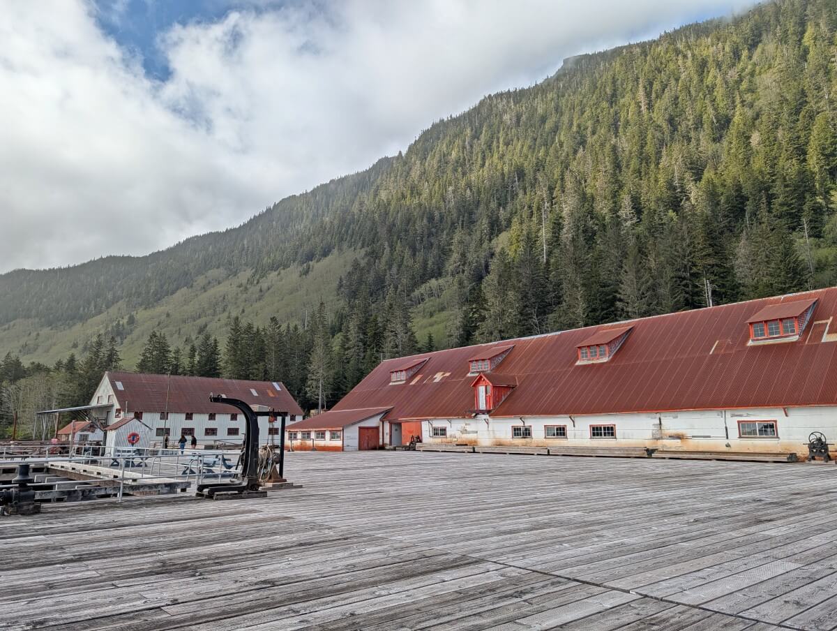 Large wooden dock area at North Pacific Cannery, in front of long red and white cannery building, backdropped by steep forested hill