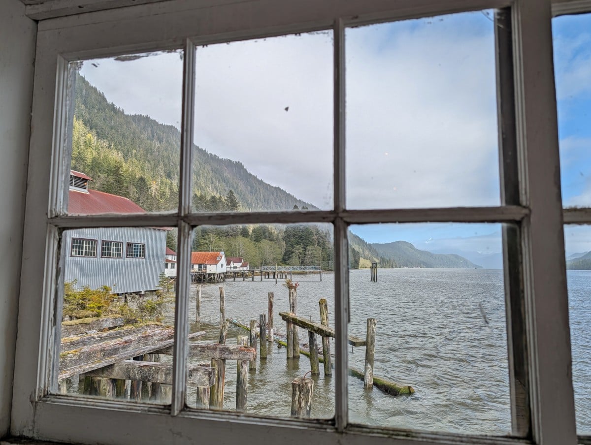 Looking through an old six panel window at the North Pacific Cannery towards pilings outside, with scattered building, calm ocean and forested hills