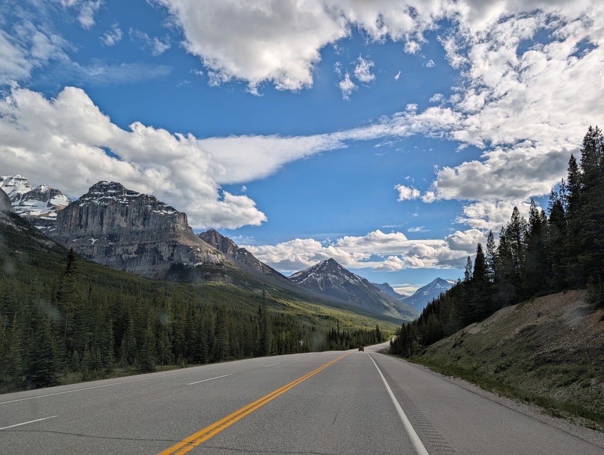 Vehicle view of Highway 93 with forested hills on side, snow capped mountains above and mountains in the distance