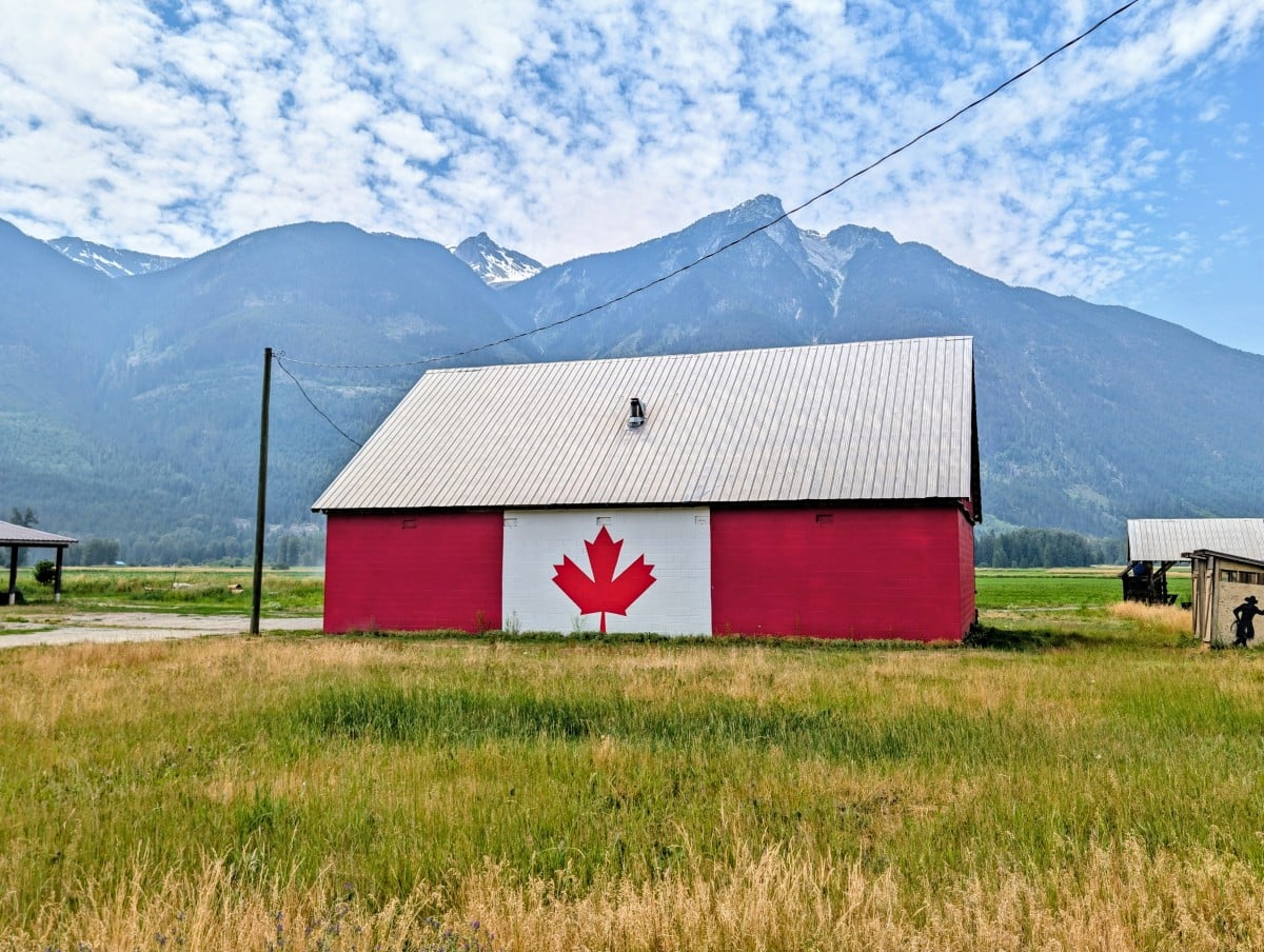 A barn sits behind a meadow, painted with the colours and maple leaf of the Canadian flag, in front of  snow capped mountains