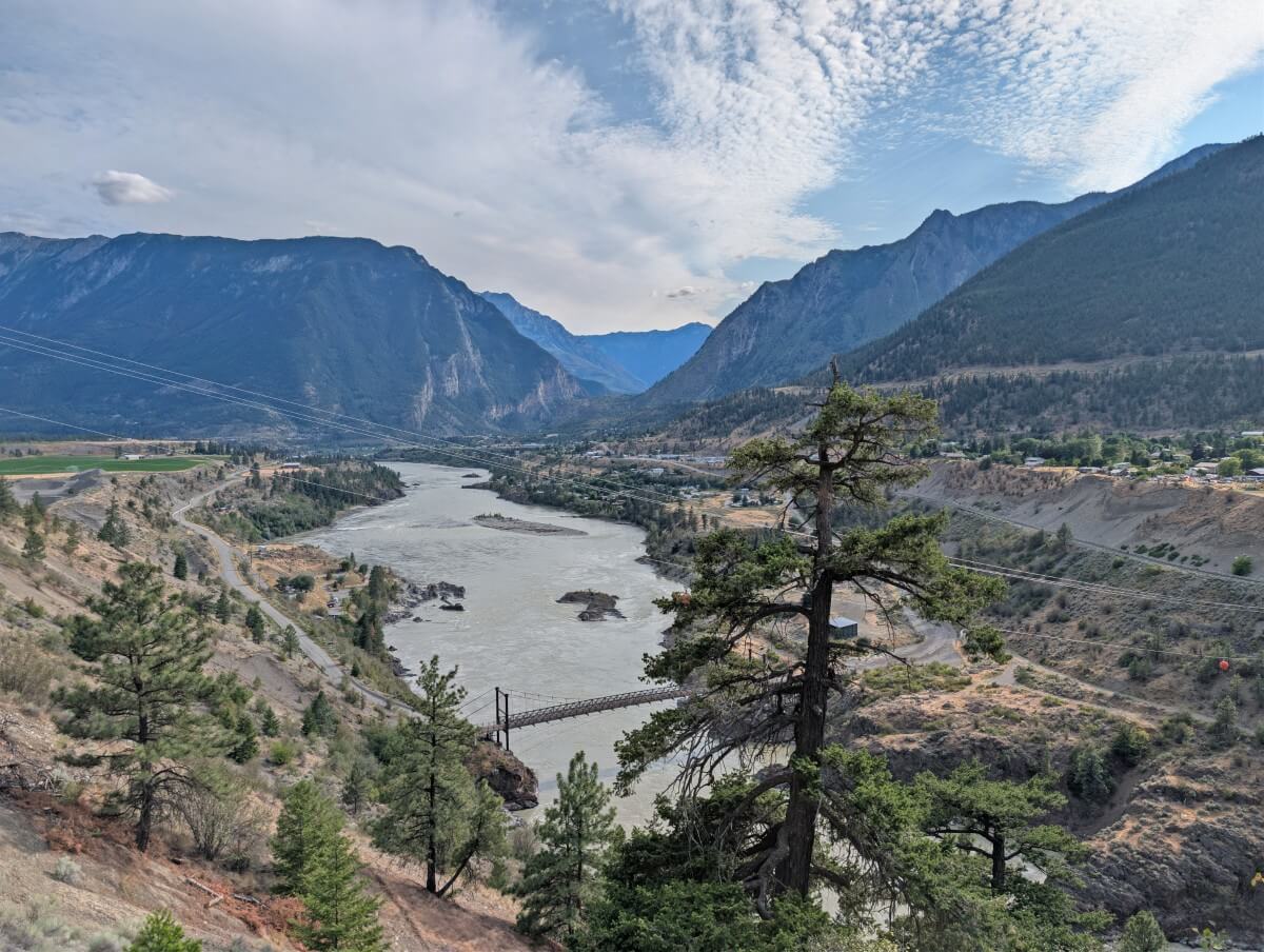 Looking over trees to the Fraser River far below, surrounded by dry slopes with scattered trees. Large mountains rise in the background, with houses above the banks of the river on the right hand side