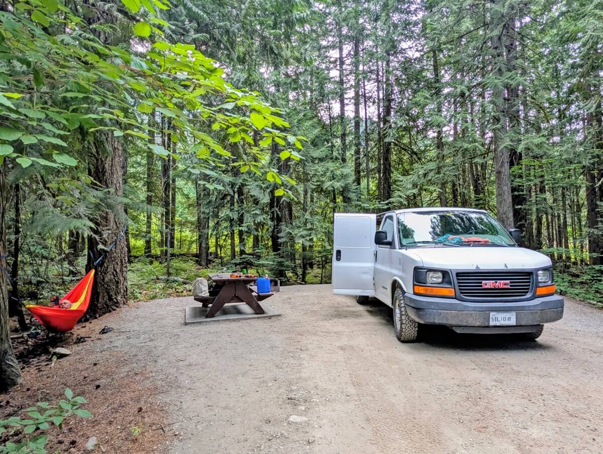 A white van is parked on a cleared camping area at Nairn Falls Provincial Park, with picnic table adjacent. There is a colourful hammock set up in the trees on the left