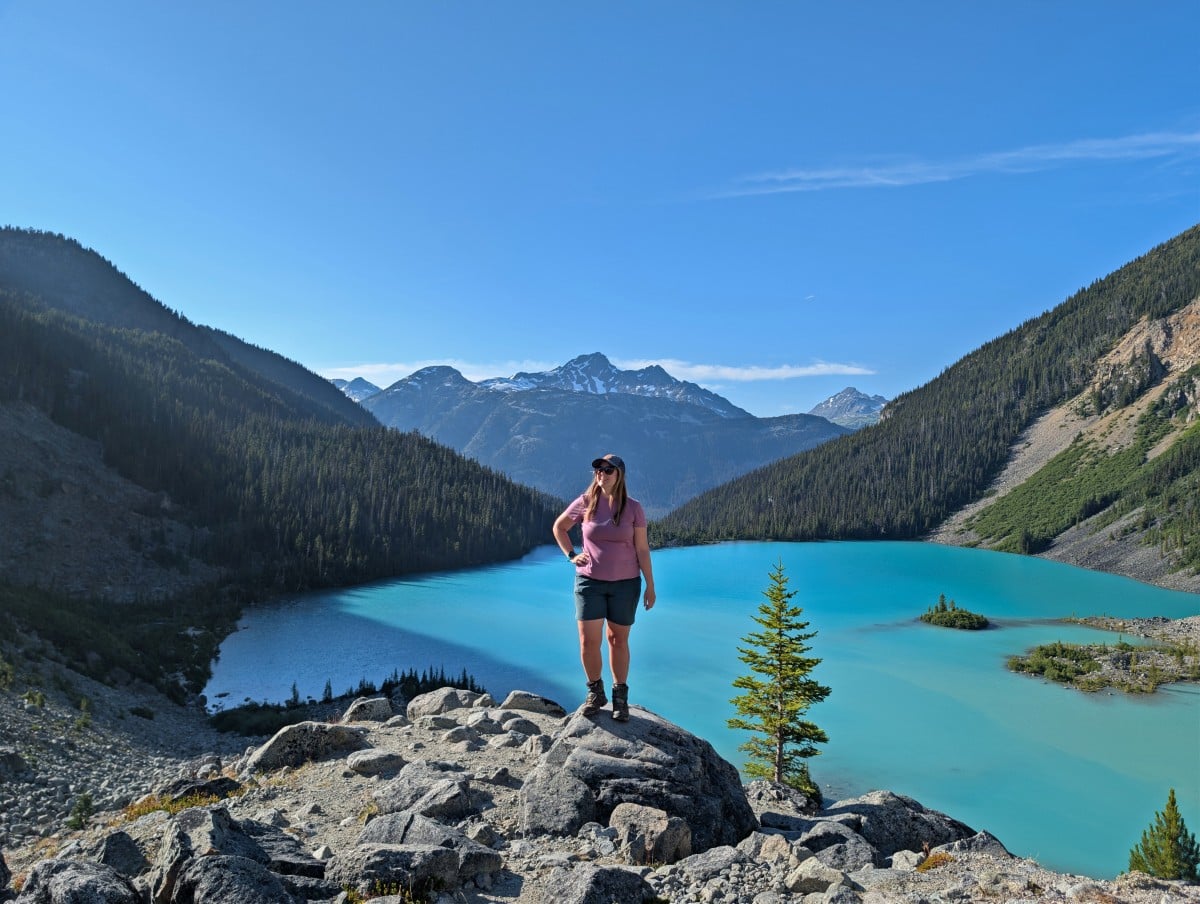 Gemma stands on a rock above turquoise coloured Joffre Lakes, with forested slopes behind, snow capped mountains in distance