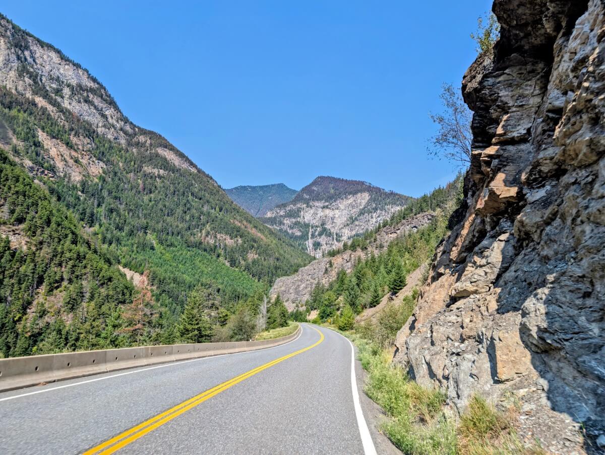 Vehicle view of Highway 99, featuring winding road with one lane on each side, rock cliff on right and forested mountains on left
