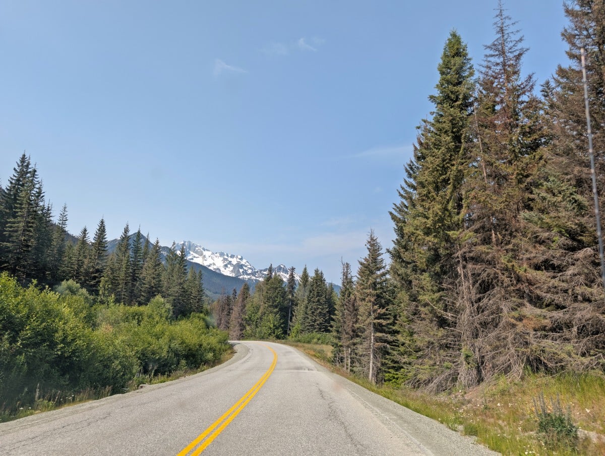 Vehicle view of Highway 99 curving around a corner to the left, lined by forest with snow capped mountains rising behind