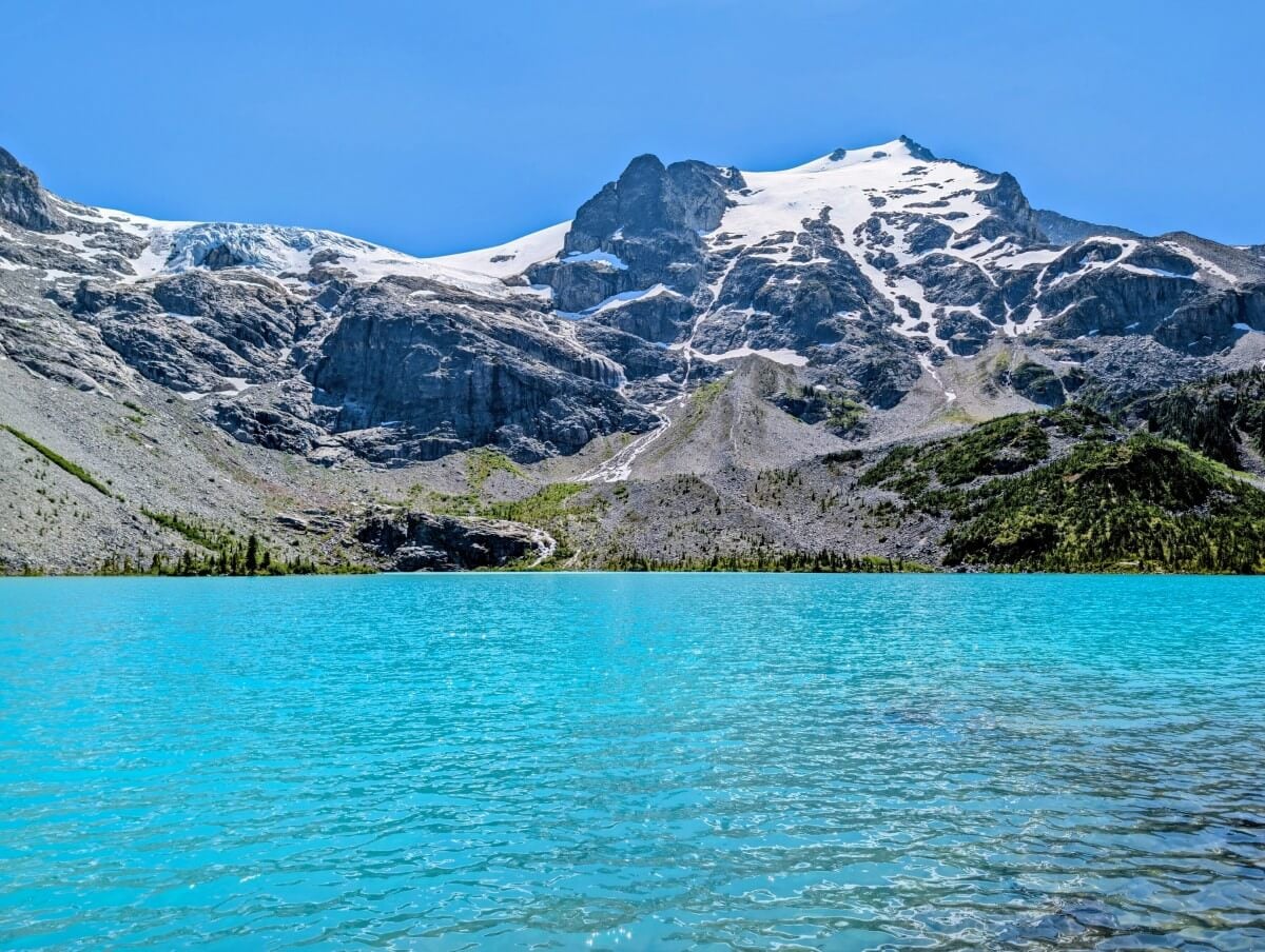 Shoreline view of Upper Joffre Lake, a bright turquoise lake backdropped by large rocky mountains, topped with a glacier