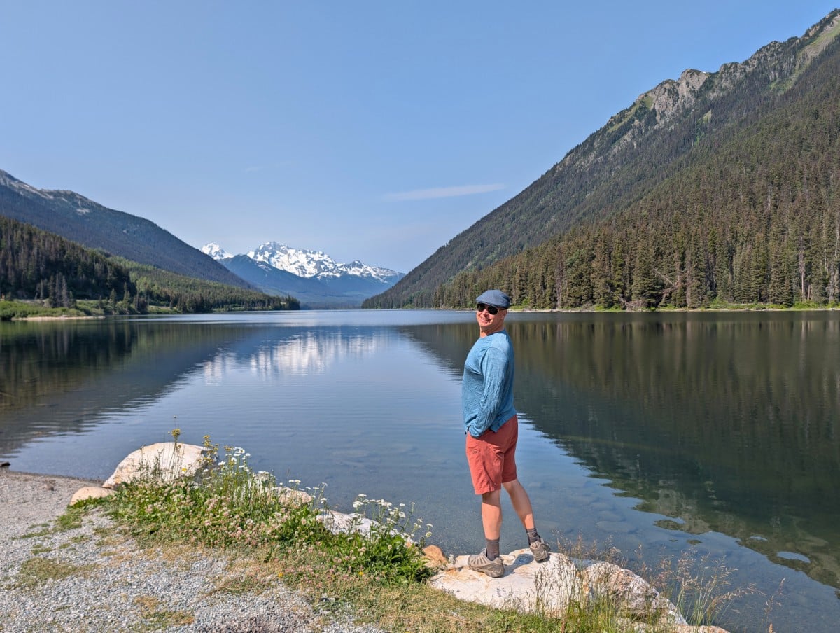 JR stands on a rock looking back at the camera, in front of calm Duffey Lake. The mountains behind the lake are reflected on the surface