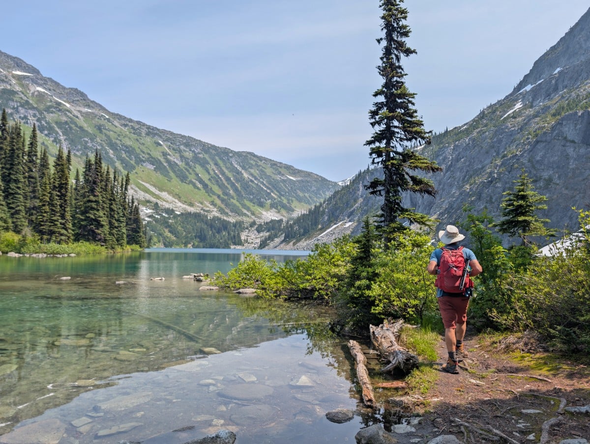 Back view of JR walking along the shoreline of a very calm clear alpine lake (Rohr Lake), backdropped by steep partially forested mountain slopes