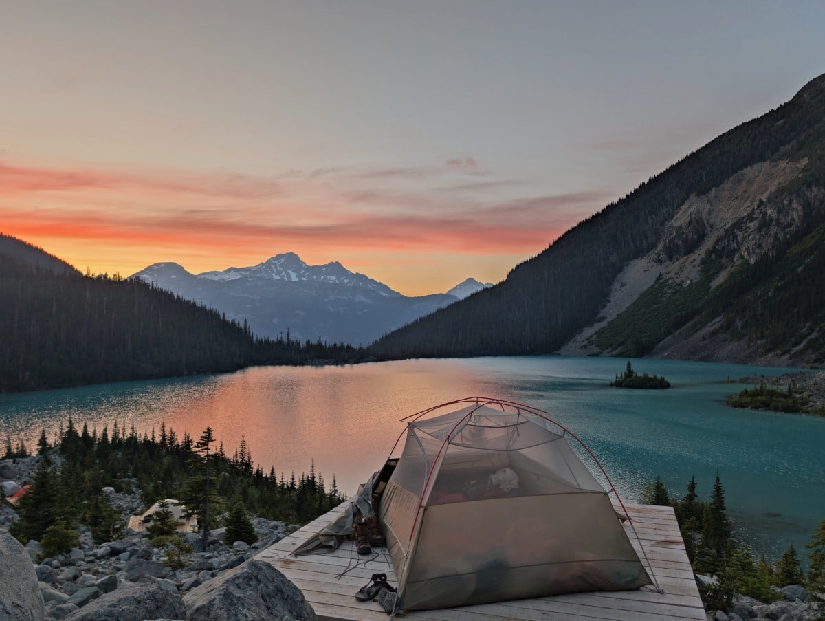 A set up tent on a wooden tent pad in front of Upper Joffre Lake at sunset, with snow capped mountains in the background
