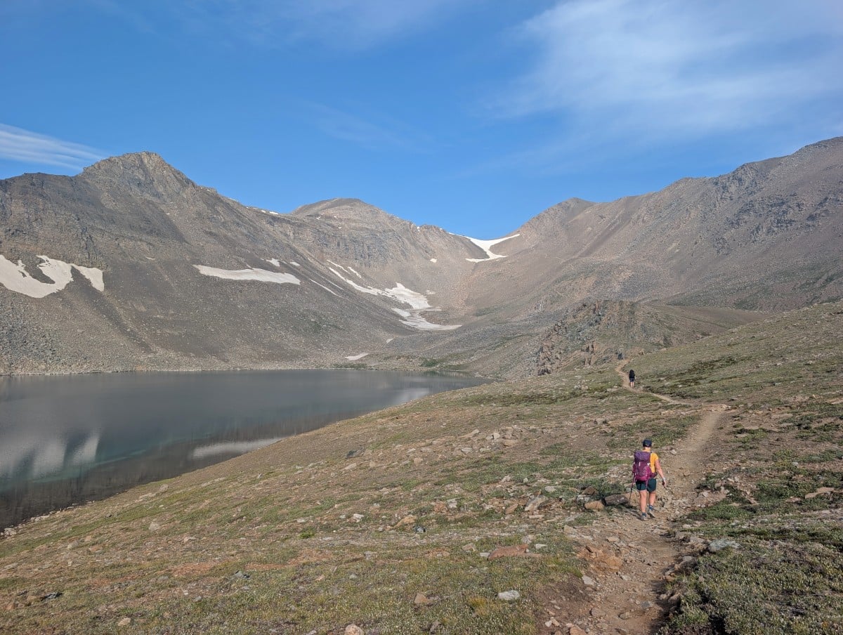 Back view of hiker walking along flat dirt path paralleling a lake, with view of a pass in the background