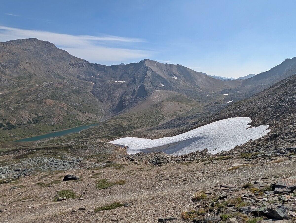 Looking across dirt trail to patch of snow on mountain slope and turquoise lake in background, backdropped by bare mountains