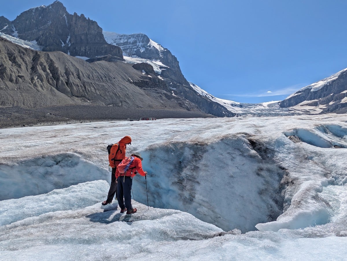 JR looks down into a glacier crevasse while standing next to tour guide, while visiting the Athabasca Glacier. There are snow capped mountains in the background