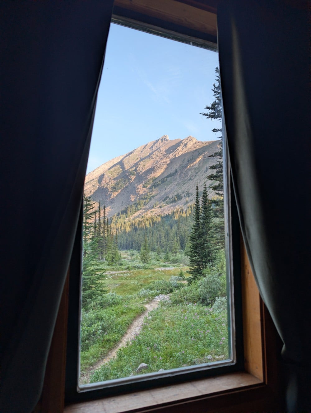 Interior cabin view looking out window (surrounded by curtains) with sloped mountain and forest outside