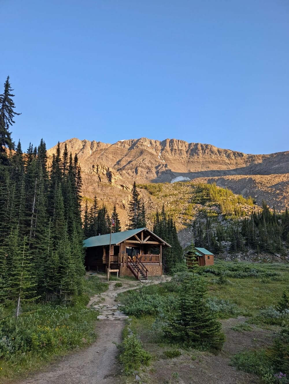 Shovel Pass Lodge, featuring main wooden lodge building with stairs, a small cabin to the right and mountains backdrop with sun shining on them
