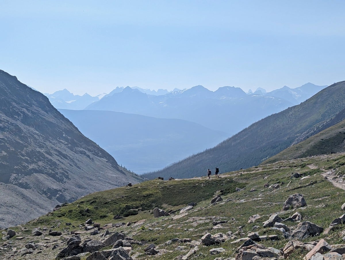 Two hikers walk down a slope in the distance with large mountains in the background