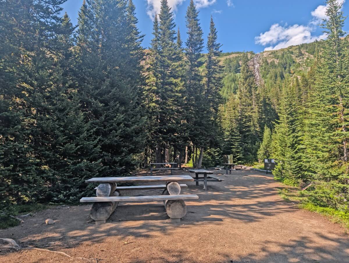 Four picnic tables set into cleared area in forest, with view of ridge and waterfall in background (Curator Campground)