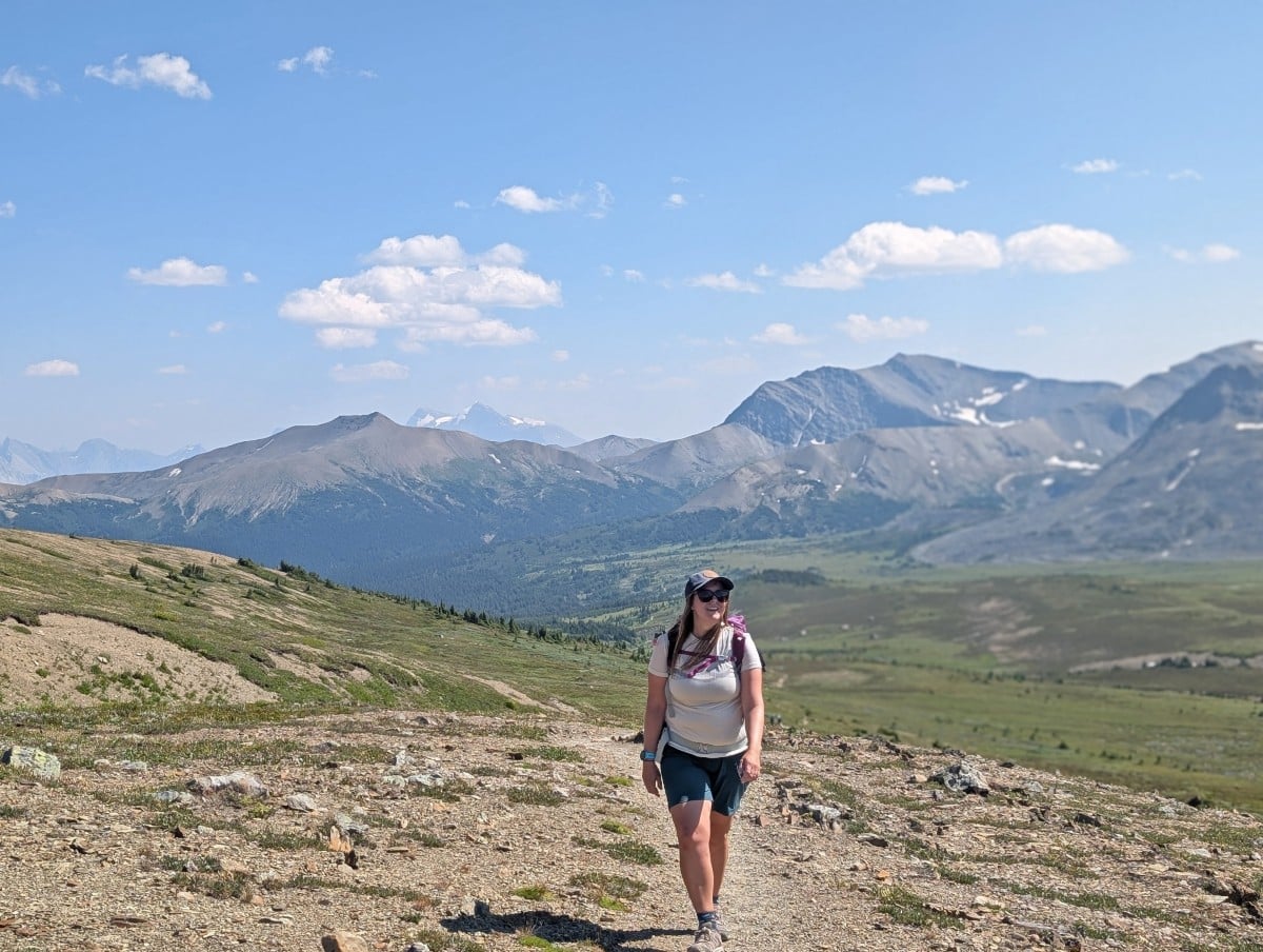 Hiker walks towards the camera on the Skyline Trail, with wide open landscape and mountains in the background