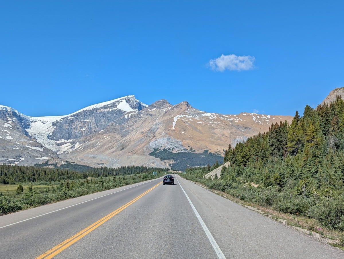 Road view of a vehicle driving on the Icefields Parkway road, lined up forest on one side and mountains on the other