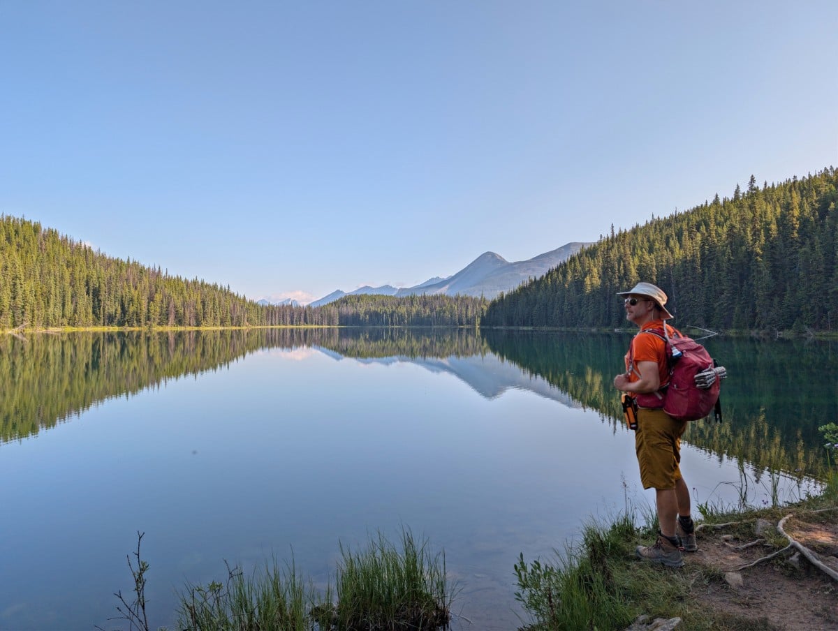 A hiker stands on the side of a very calm lake, which is reflected the trees on the shoreline and mountains in the background