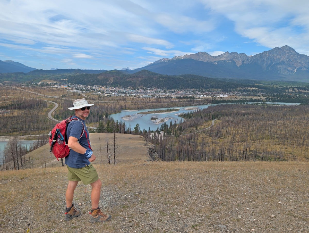 Side view of JR standing at Old Fort Point viewpoint, with milky turquoise Athabasca River in the background with Jasper Townsite, backdropped by mountains