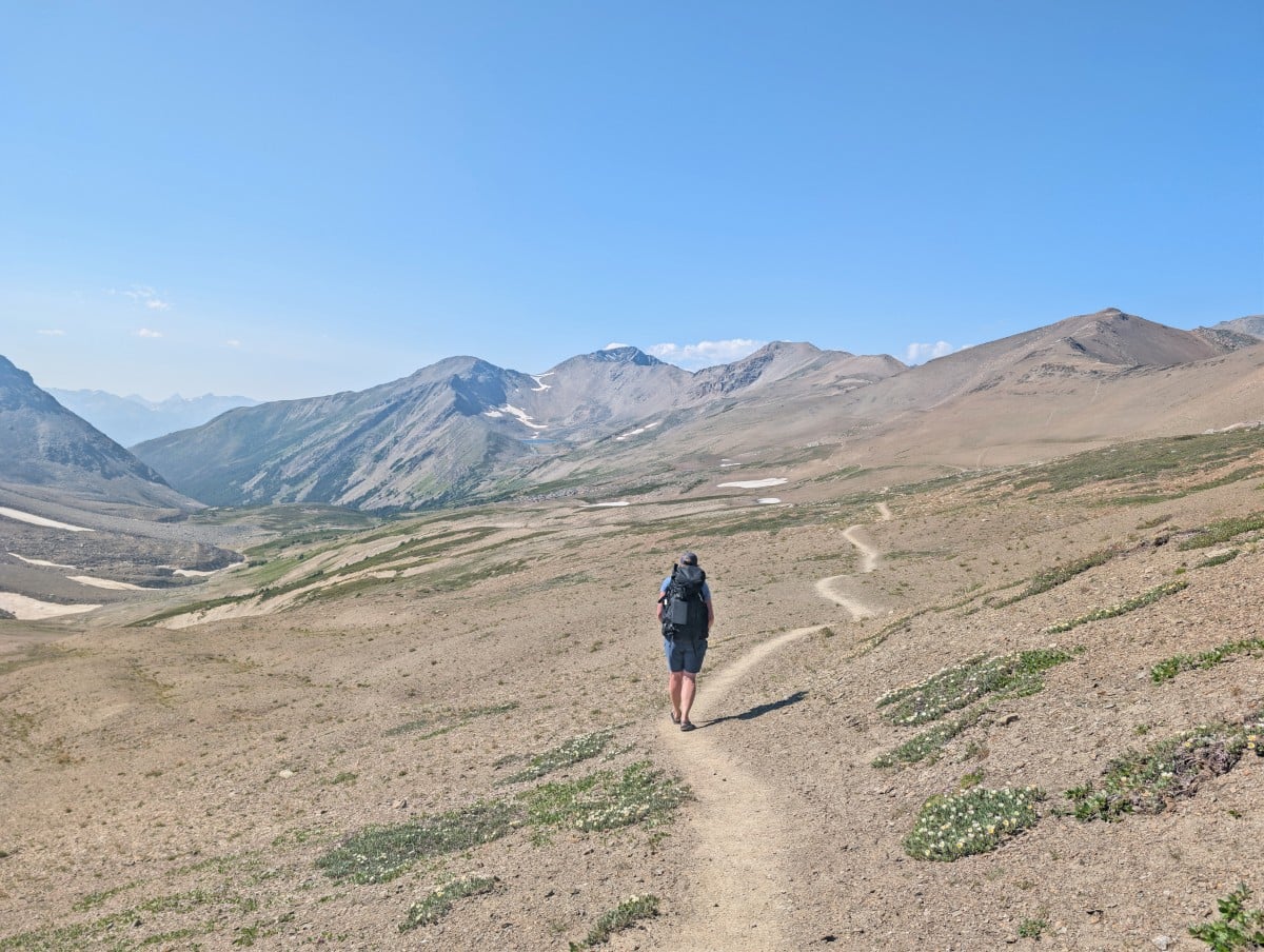 Back view of Leigh hiking with a backpack along a dirt trail through a very open alpine ladnscape backdropped by mountains