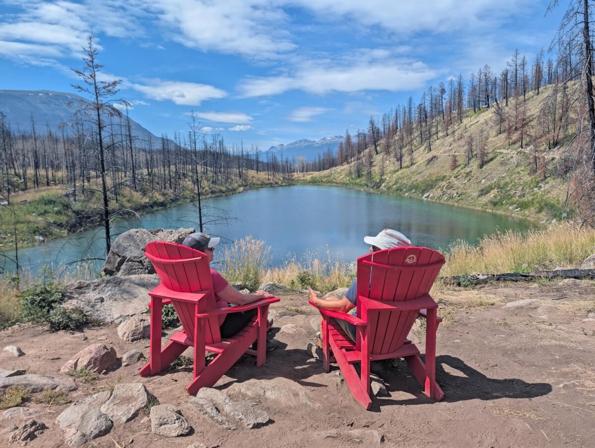 Back view of two people sitting in red Adirondack chairs in front of a turquoise lake