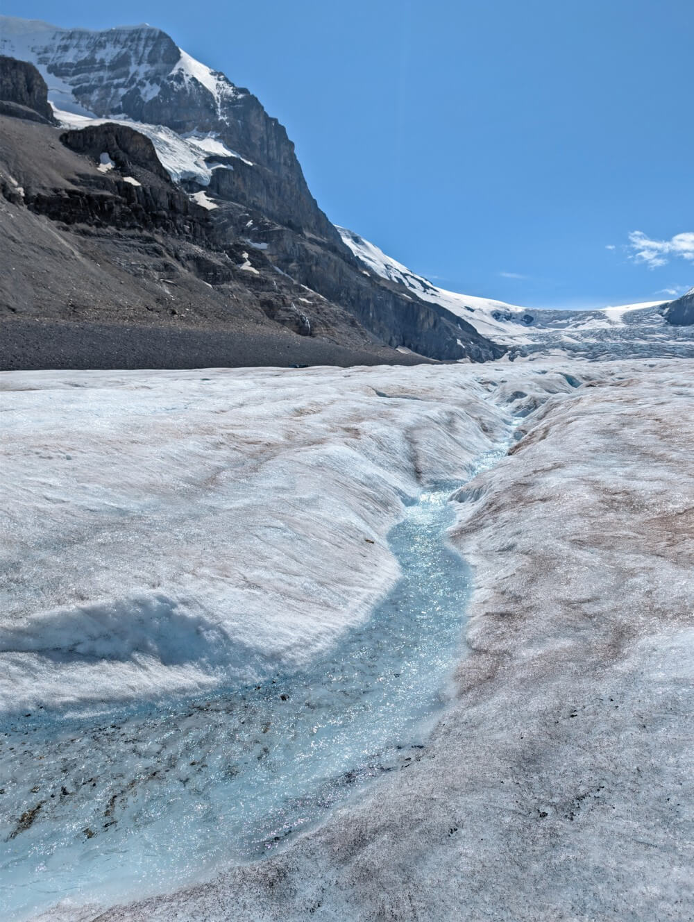 A channel of water runs through glacial ice at the Athabasca Glacier with mountains and main part of glacier in the background