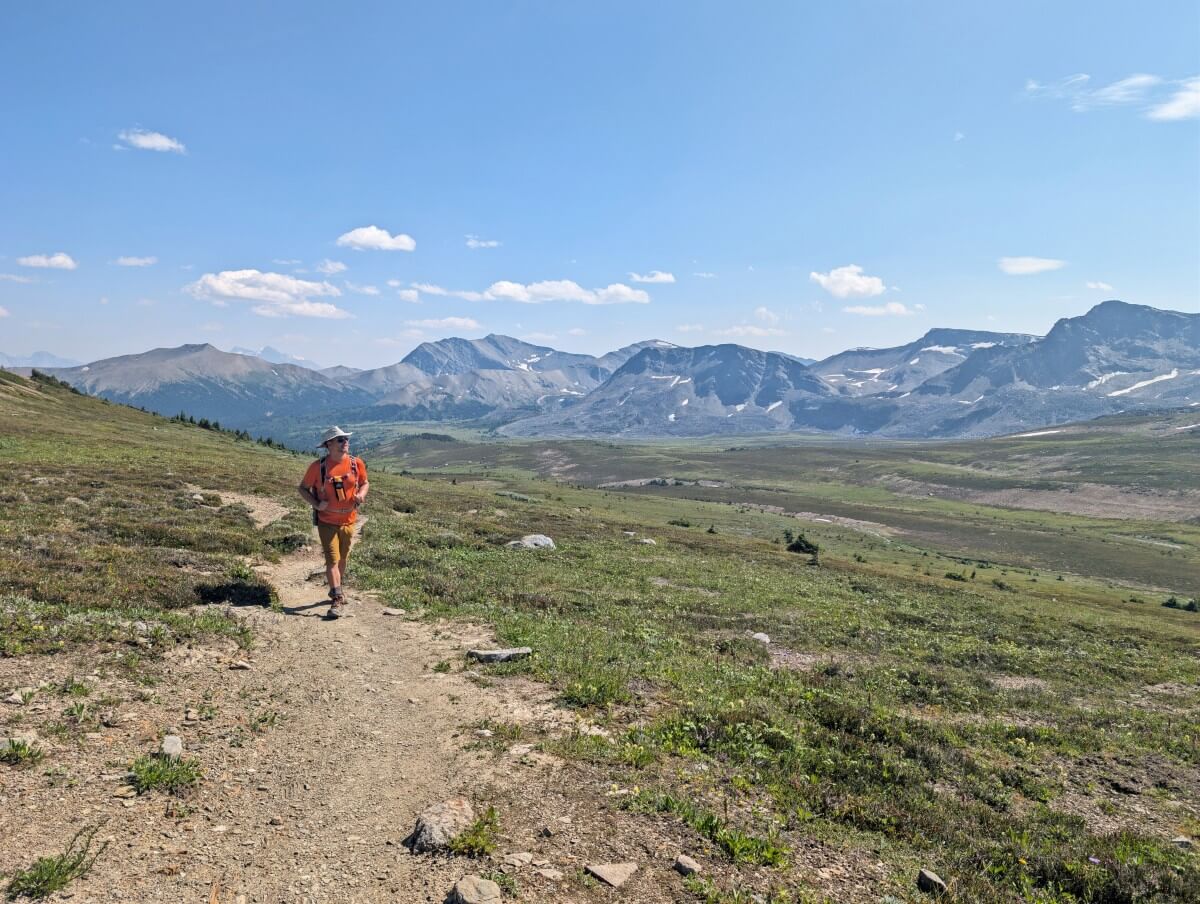 A hiker walks towards the camera on a dirt path with meadows and mountains in the background