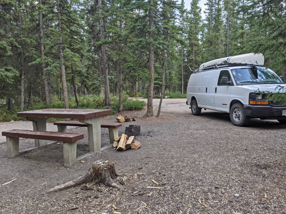 A white van with canoe on top is parked on gravel tent pad in the forest at Snaring Campground, with picnic table and fire pit close to camera