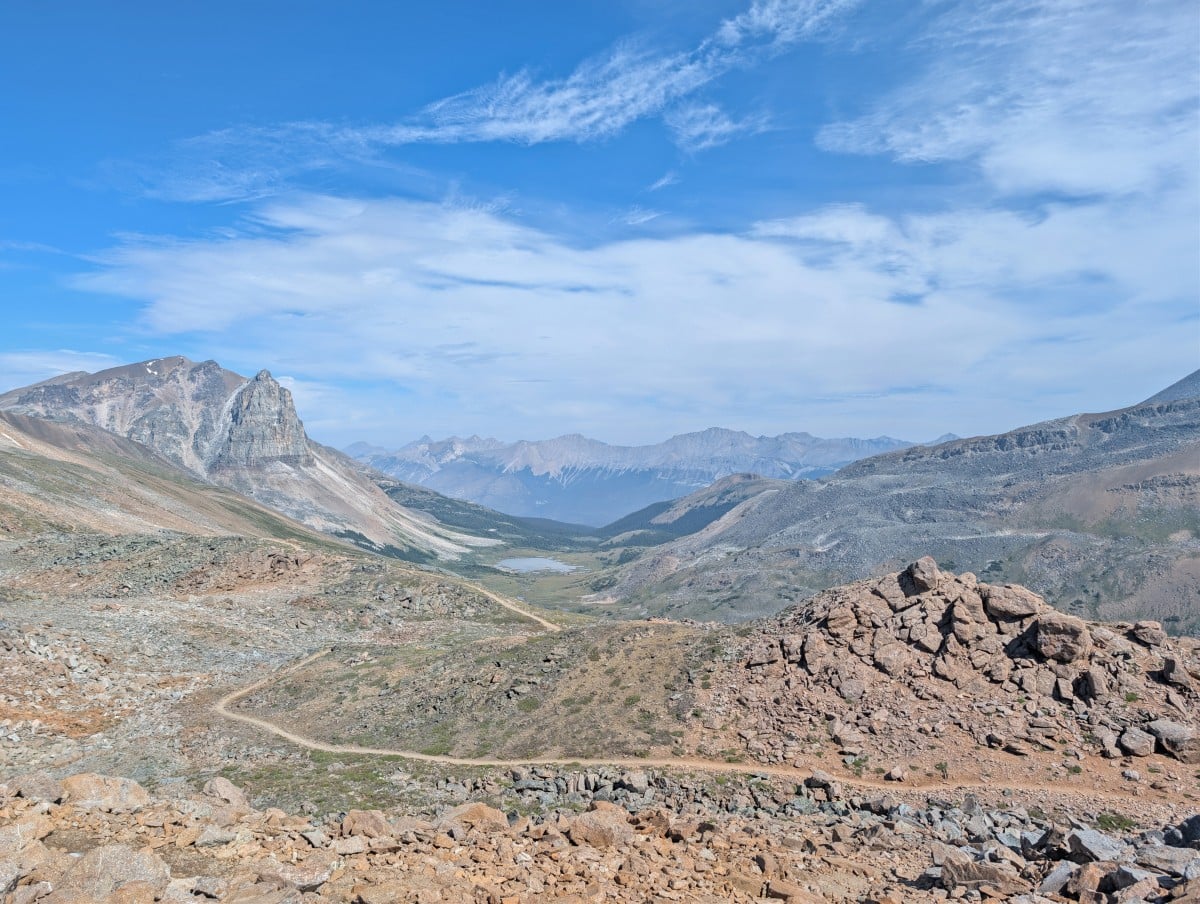 A dirt trail leads across and down into a valley with a lake, framed by mountains