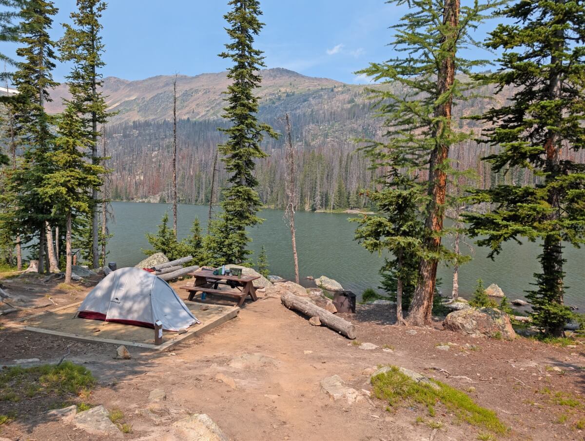 Looking down to set up tent on dirt wooden framed tent pad next to Quiniscoe Lake with picnic table and scattered trees