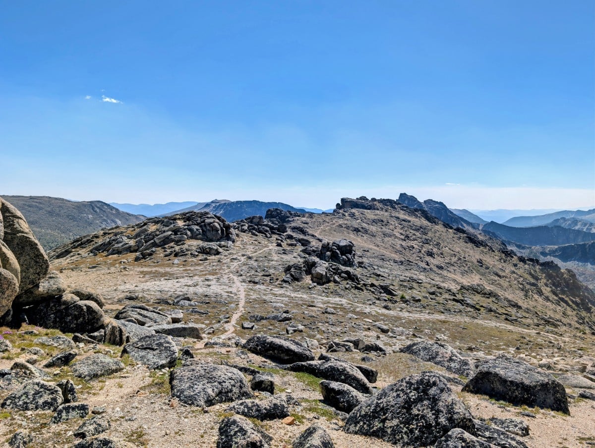 A dirt path snakes through open alpine terrain with scattered rocks and mountain layers in background