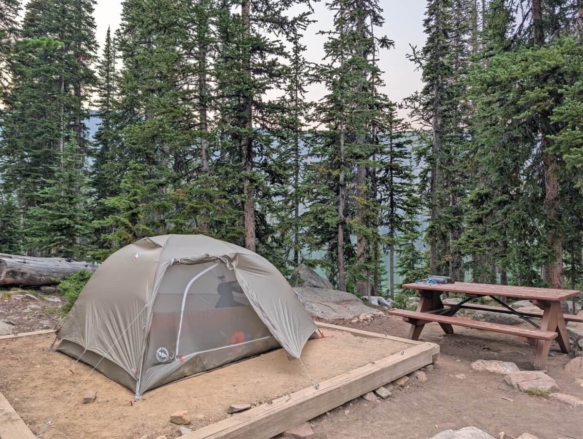 A set up tent on dirt tent pad next to picnic table, backdropped by trees, with lake visible through them at Quiniscoe Lake Campground