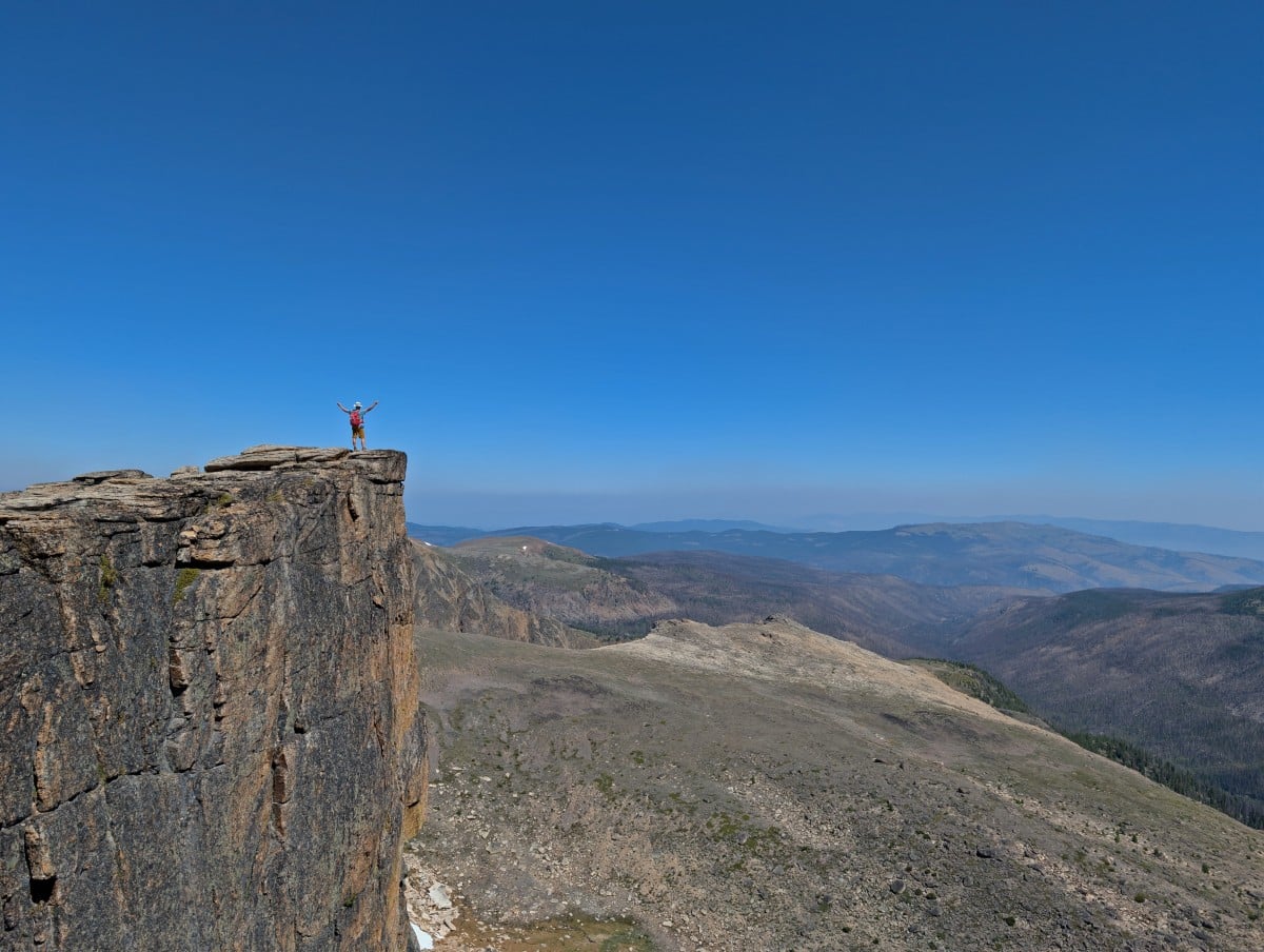 A man stands on a rock formation with a vertical edge with arms outstretched, looking out to rounded mountain layers